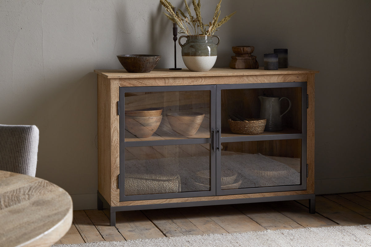 The nkuku Nirvi Mango Wood & Iron Sideboard - Black features glass doors showcasing bowls, baskets, and linens. On top sit a vase with dried flowers and bowls. The sideboard stands on a wooden floor against a neutral wall.