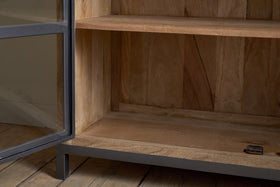 A close-up of the nkuku Nirvi Mango Wood & Iron Sideboard in black, featuring a metal frame, open glass door, and two shelves on a wooden floor. A small metal object sits on the bottom shelf.