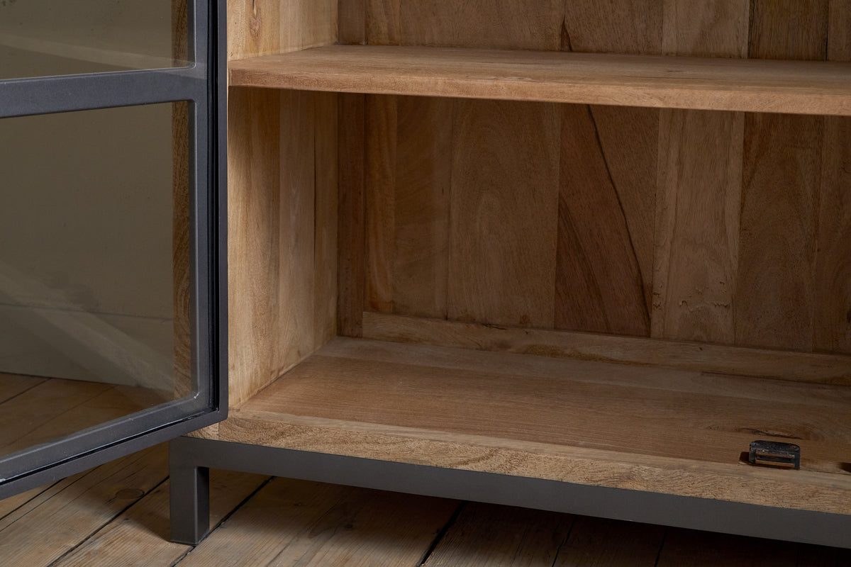 A close-up of the nkuku Nirvi Mango Wood & Iron Sideboard in black, featuring a metal frame, open glass door, and two shelves on a wooden floor. A small metal object sits on the bottom shelf.