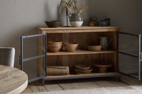 The nkuku Nirvi Mango Wood & Iron Sideboard - Black features black metal-framed glass doors that open to display wooden bowls, a pitcher, and baskets inside, while pottery and dried grasses decorate the top.