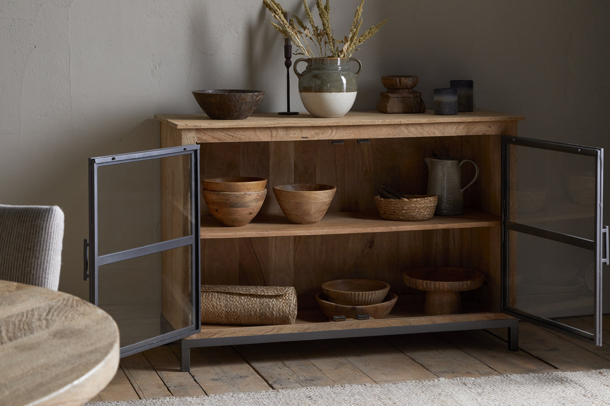 The nkuku Nirvi Mango Wood & Iron Sideboard - Black features black metal-framed glass doors that open to display wooden bowls, a pitcher, and baskets inside, while pottery and dried grasses decorate the top.