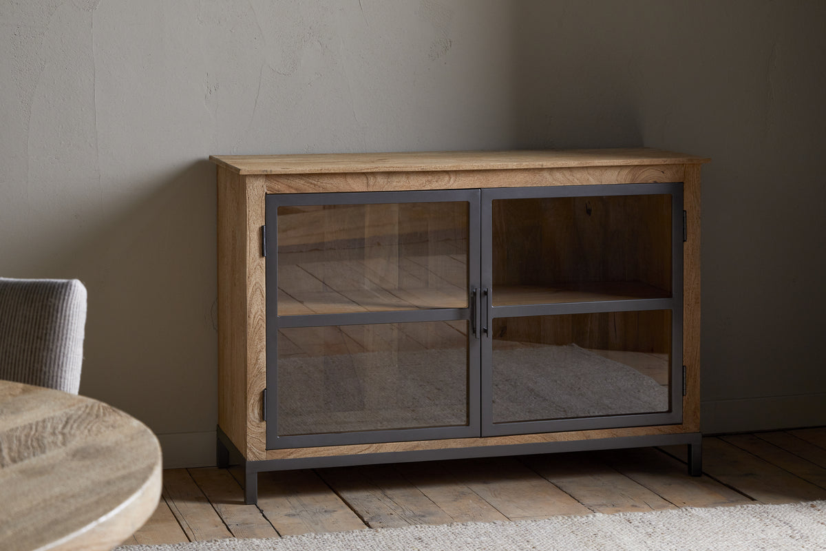 The nkuku Nirvi Mango Wood & Iron Sideboard - Black features glass-front double doors and a black frame, standing on a wooden floor against a light wall in a minimalist room with part of a round table and upholstered chair visible on the left.