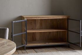 The Nirvi Mango Wood & Iron Sideboard by nkuku, featuring glass doors and a single shelf, sits open on a wooden floor beside a round table and cushioned chair in a softly lit room with textured walls.