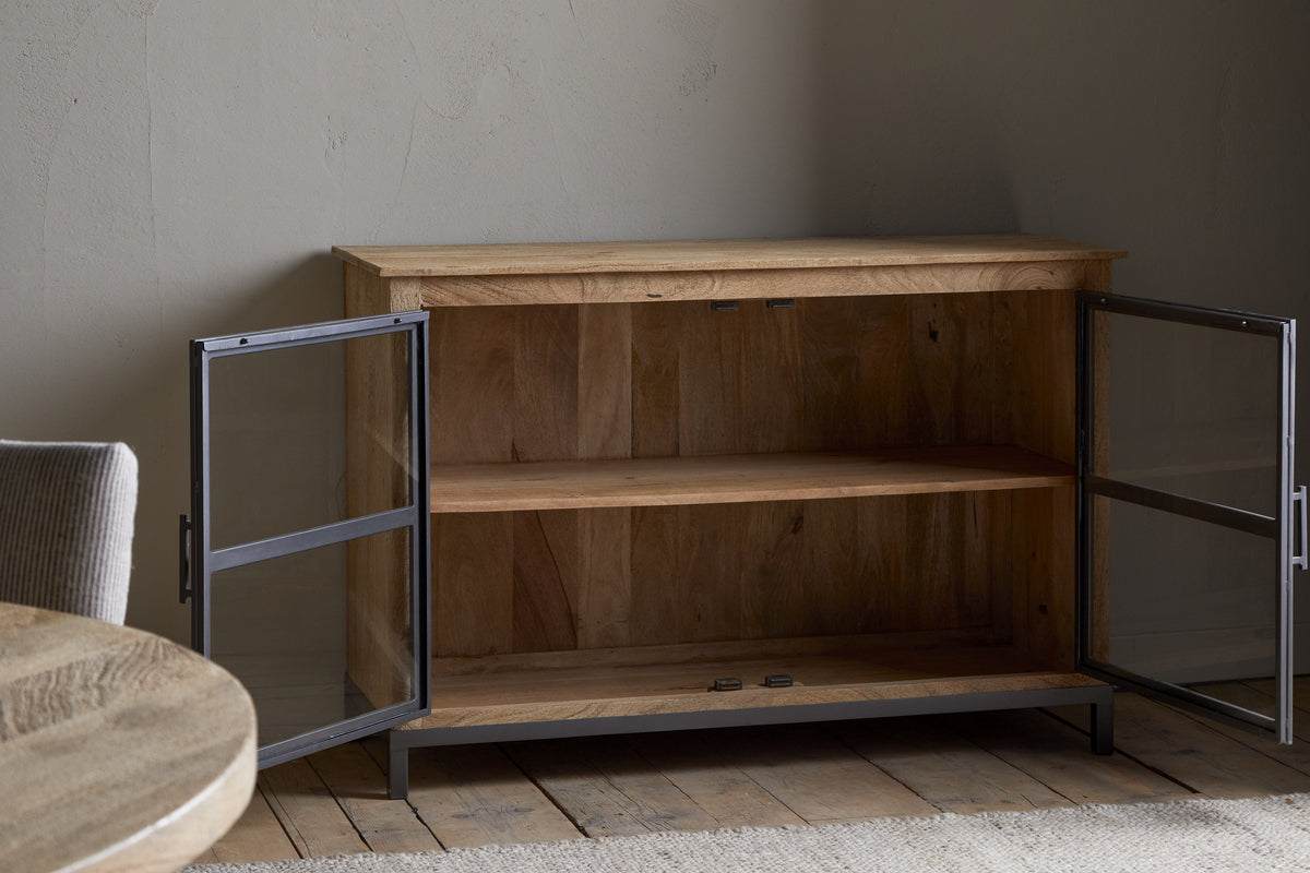 The Nirvi Mango Wood & Iron Sideboard by nkuku, featuring glass doors and a single shelf, sits open on a wooden floor beside a round table and cushioned chair in a softly lit room with textured walls.