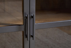 Close-up of two black metal handles on the glass cabinet doors of the nkuku Nirvi Mango Wood & Iron Sideboard - Black, showcasing the detailed texture of its iron and mango wood surfaces and wooden interior shelves.