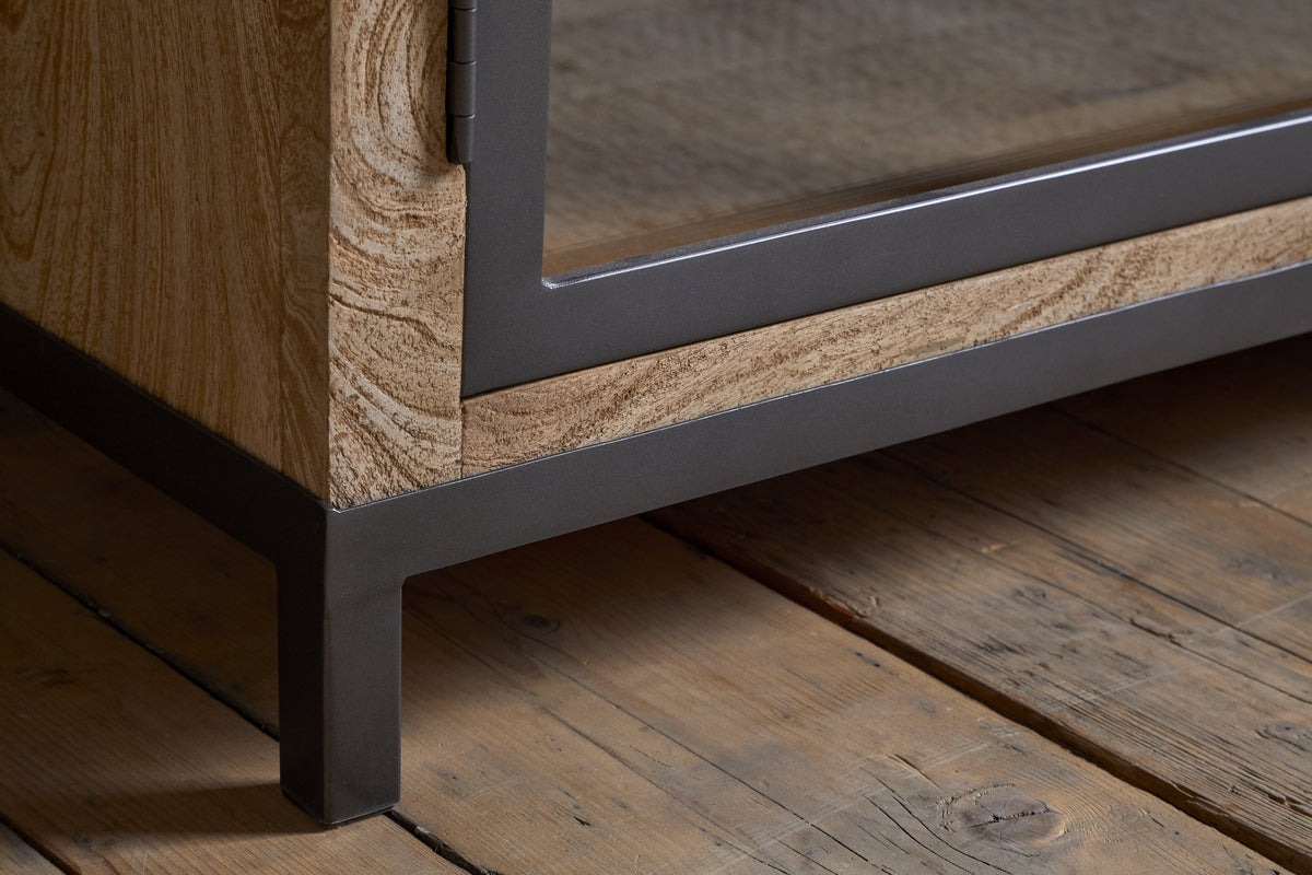Close-up of the Nirvi Mango Wood & Iron Sideboard - Black by nkuku, showcasing its light mango wood grain, black iron frame, and glass panel door, set on a rustic wooden floor with natural texture.