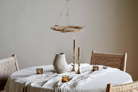 A round table with a cream cloth features a ribbed ceramic vase, brass candle holders, and brown votives. Two woven chairs sit nearby, all beneath the elegant nkuku Natesa Hanging Wreath - Brass and a rustic twig chandelier overhead.