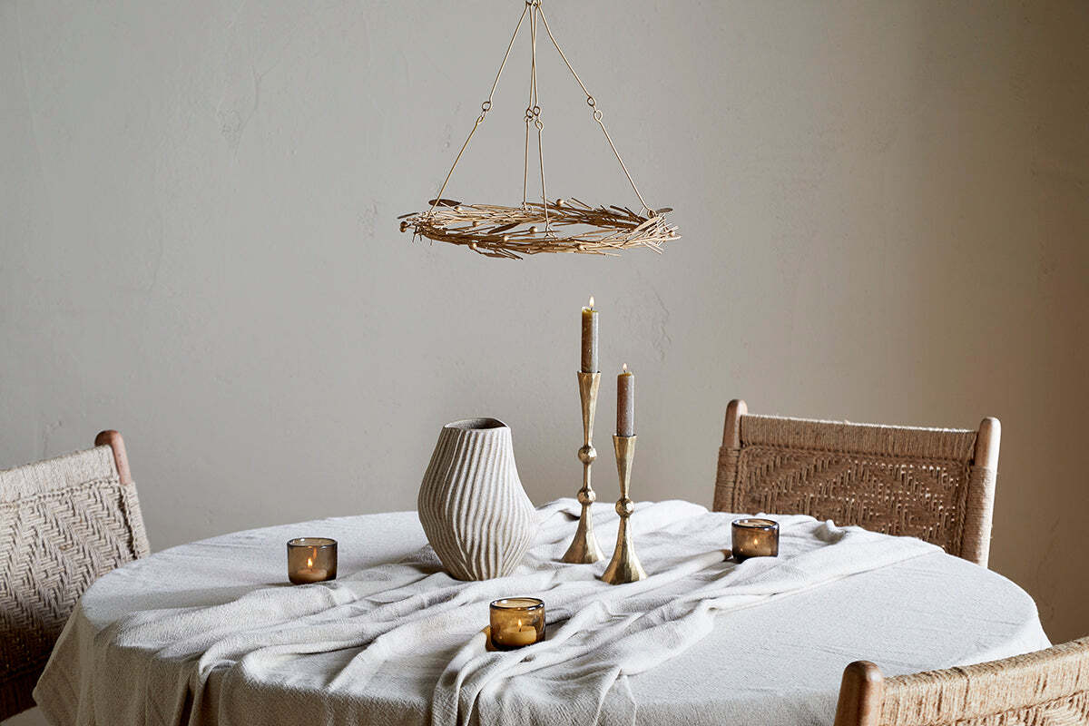 A round table with a cream cloth features a ribbed ceramic vase, brass candle holders, and brown votives. Two woven chairs sit nearby, all beneath the elegant nkuku Natesa Hanging Wreath - Brass and a rustic twig chandelier overhead.