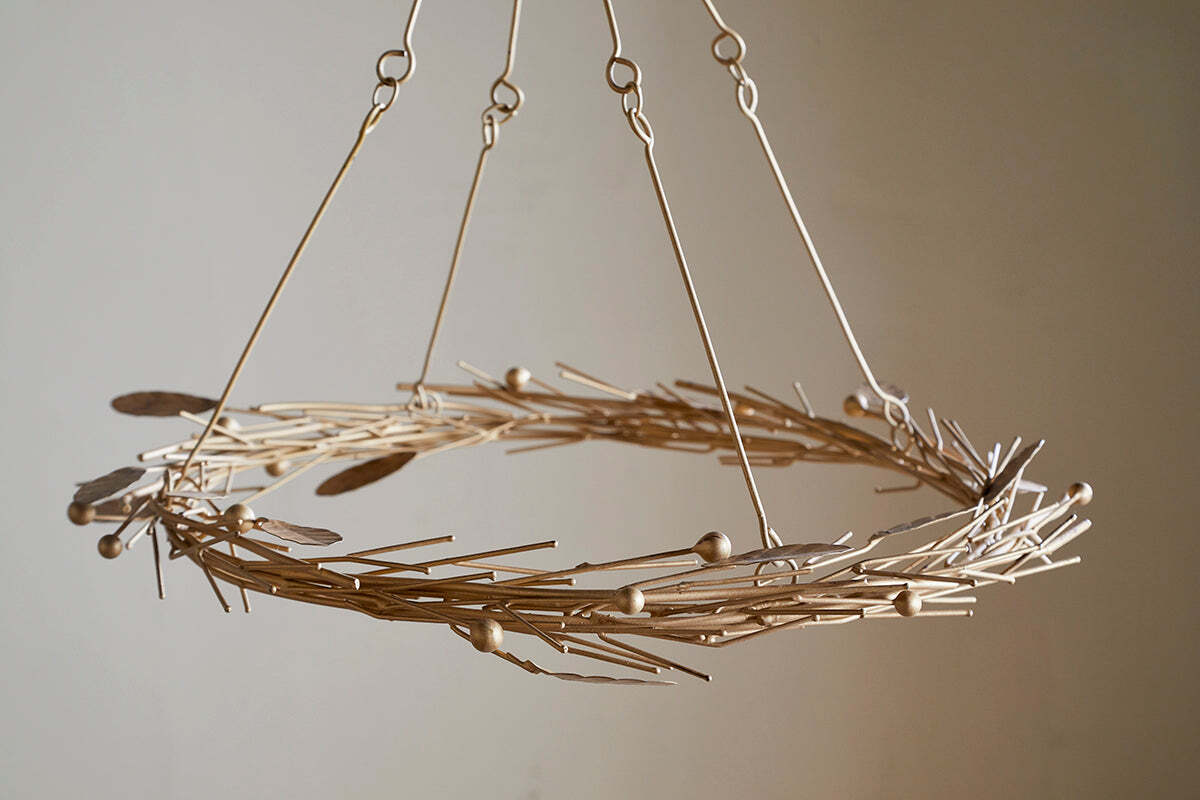 The Natesa Hanging Wreath - Brass by nkuku is a modern chandelier featuring thin gold rods, small spheres, and feather-shaped pieces. Suspended with four metal rods, it stands out against a plain beige background.
