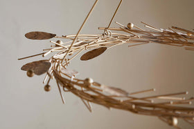 A close-up of the Natesa Hanging Wreath - Brass by nkuku showcases an abstract, circular sculpture crafted from thin brass rods, oval shapes, and small spheres. Its intertwined form evokes elegant traditional craftsmanship against a softly blurred background.