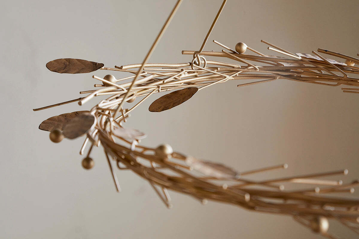 A close-up of the Natesa Hanging Wreath - Brass by nkuku showcases an abstract, circular sculpture crafted from thin brass rods, oval shapes, and small spheres. Its intertwined form evokes elegant traditional craftsmanship against a softly blurred background.