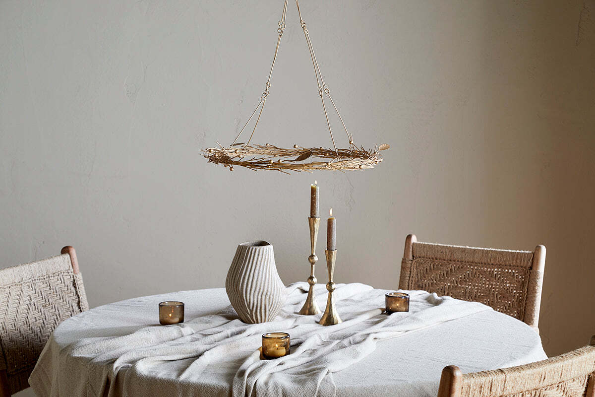 A round table with a white textured cloth displays a ribbed ceramic vase, brass candlesticks, and brown glass tealight holders. Above, the nkuku Natesa Hanging Wreath - Brass chandelier adds elegance against a plain beige wall.
