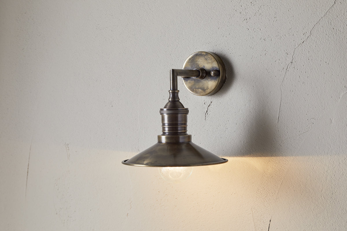 The Najja Brass Wall Lamp by nkuku features an antique brass metal shade and exposed bulb, mounted on a light-textured wall to emit a warm, soft glow with timeless elegance.