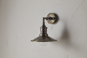 The Najja Brass Wall Lamp by nkuku features a vintage-style wide, shallow shade and exposed bulb in an antique brass finish, elegantly mounted on a light-colored, textured wall.