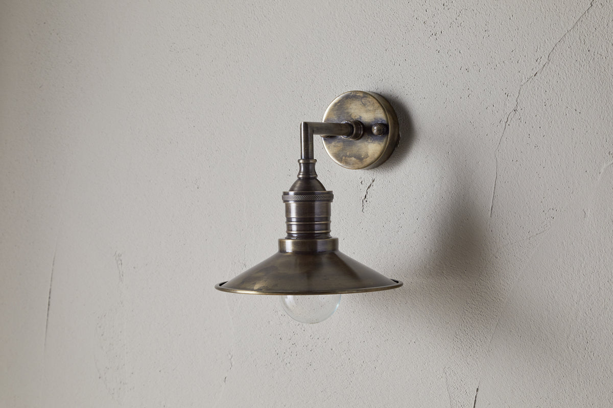 The Najja Brass Wall Lamp by nkuku features a vintage-style wide, shallow shade and exposed bulb in an antique brass finish, elegantly mounted on a light-colored, textured wall.