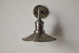 The Najja Brass Wall Lamp by nkuku features a vintage-style, wide shallow metal shade and exposed bulb in an antique brass finish, mounted on a light-colored textured wall.