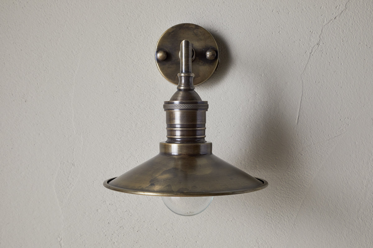 The Najja Brass Wall Lamp by nkuku features a vintage-style, wide shallow metal shade and exposed bulb in an antique brass finish, mounted on a light-colored textured wall.
