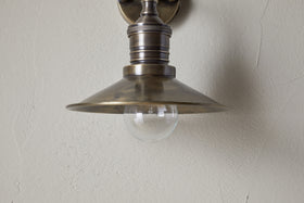 A close-up of the nkuku Najja Brass Wall Lamp in antique brass, featuring a metal shade and exposed clear bulb, mounted on a light-colored textured wall.