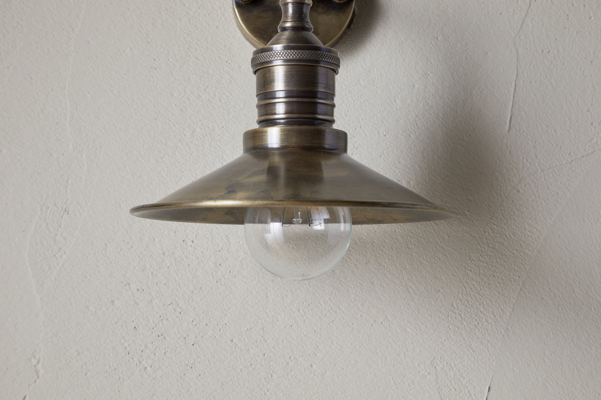 A close-up of the nkuku Najja Brass Wall Lamp in antique brass, featuring a metal shade and exposed clear bulb, mounted on a light-colored textured wall.
