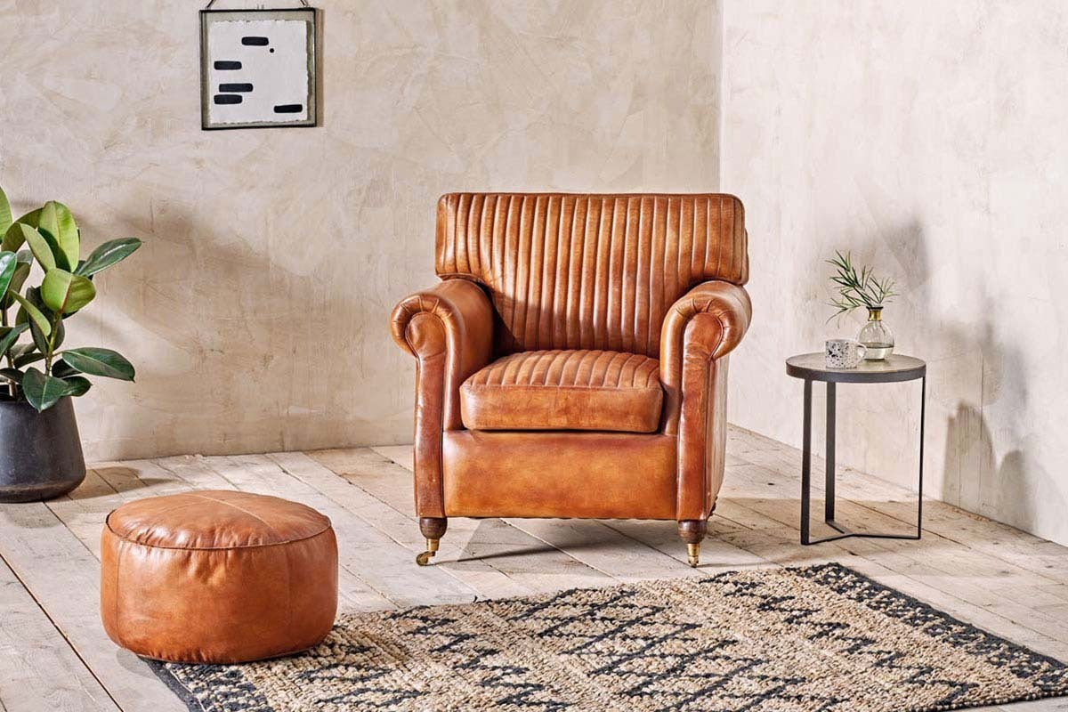 A cozy room features a vintage brown leather armchair with vertical stitching, the nkuku Nadu Leather Pouf in Aged Tan for extra seating or as a footrest, a small black side table with décor, leafy greenery in a black pot, wall art, and a patterned beige rug.