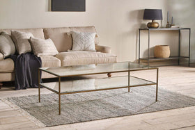 A modern living room with a beige sofa, neutral throw pillows, and the nkuku Nakuru Iron & Glass Coffee Table on a gray rug. A console table with decor and a woven basket stands against a light wall, all atop light wood flooring.