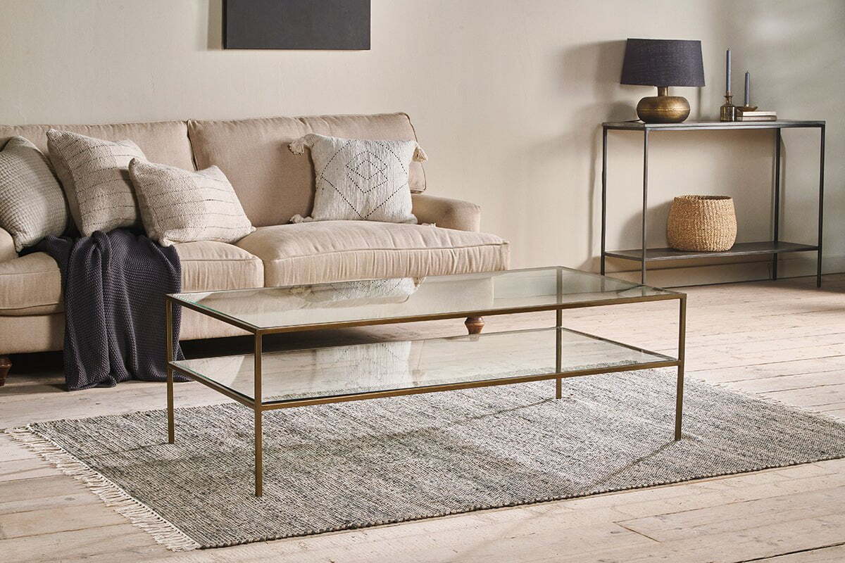 A modern living room with a beige sofa, neutral throw pillows, and the nkuku Nakuru Iron & Glass Coffee Table on a gray rug. A console table with decor and a woven basket stands against a light wall, all atop light wood flooring.