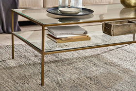 A nkuku Nakuru Iron & Glass Coffee Table with a gold frame stands on a textured gray rug. Its glass shelf holds books and a wooden box, while the tabletop displays a round tray, glass vase, and decor. A beige sofa is in the background.