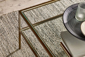 A nkuku Nakuru Iron & Glass Coffee Table stands on a textured woven carpet. On the table are stacked books, a dark patterned plate, and a ribbed glass tumbler. A wooden floor is visible in the upper left corner.
