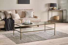 A modern living room with a beige sofa, textured pillows, dark throw blanket, and the nkuku Nakuru Iron & Glass Coffee Table with a slim gold frame and glass shelf on a woven rug, plus a console table with decor and basket against a neutral wall.