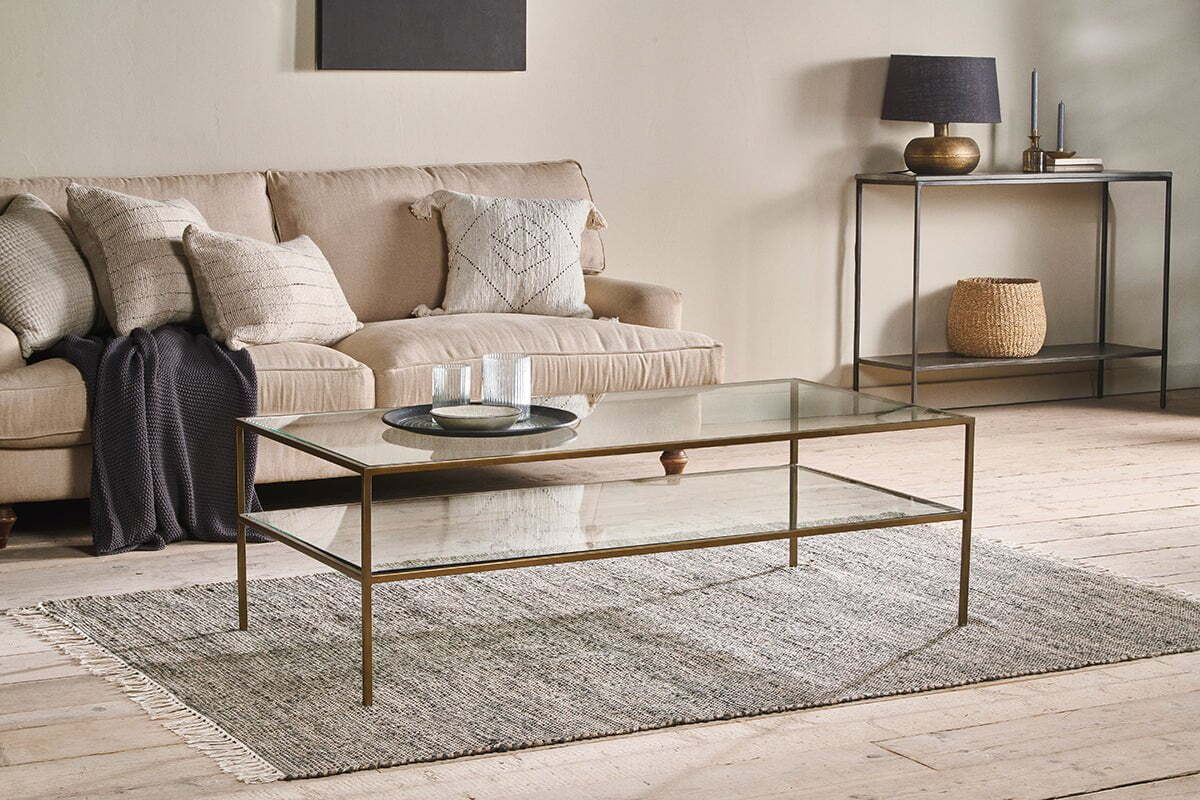 A modern living room with a beige sofa, textured pillows, dark throw blanket, and the nkuku Nakuru Iron & Glass Coffee Table with a slim gold frame and glass shelf on a woven rug, plus a console table with decor and basket against a neutral wall.