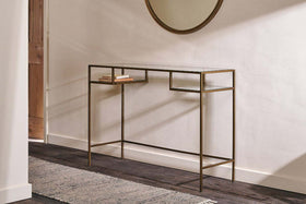 The Nkuku Nakuru Iron & Glass Console Table stands against a white wall, displaying two stacked books. A round mirror hangs above, while natural light from the left highlights the dark wood floor and textured rug.