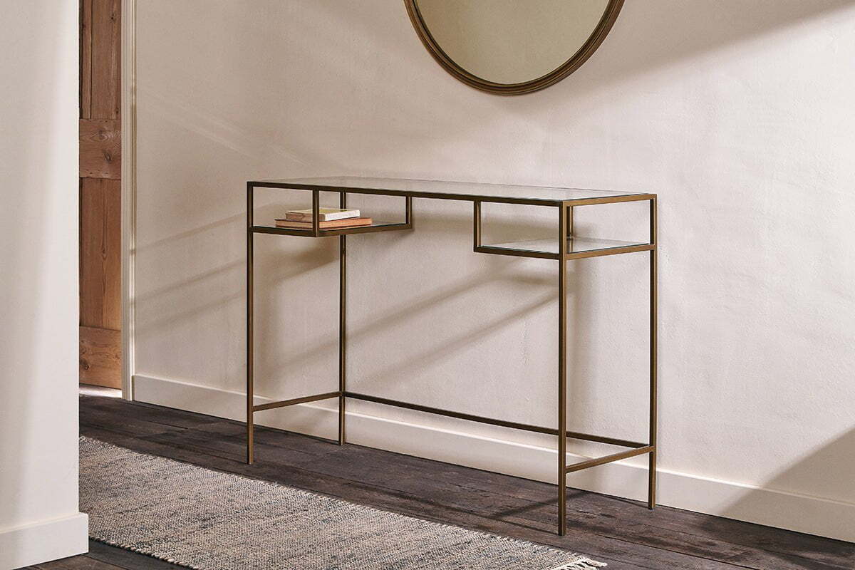The Nkuku Nakuru Iron & Glass Console Table stands against a white wall, displaying two stacked books. A round mirror hangs above, while natural light from the left highlights the dark wood floor and textured rug.