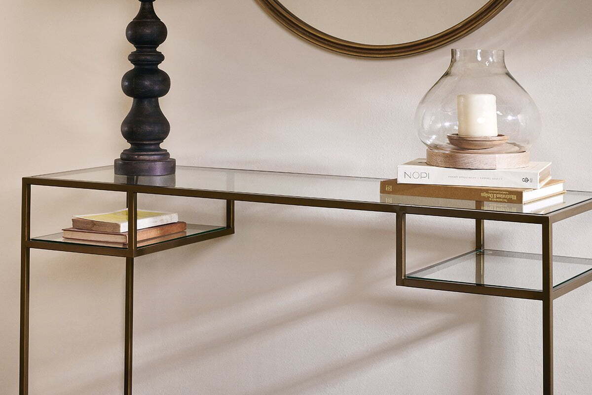 The nkuku Nakuru Iron & Glass Console Table, topped with stacked books, a black lamp, and a glass hurricane vase with a white candle, sits beneath a round mirror that reflects the table’s elegant, contemporary style.
