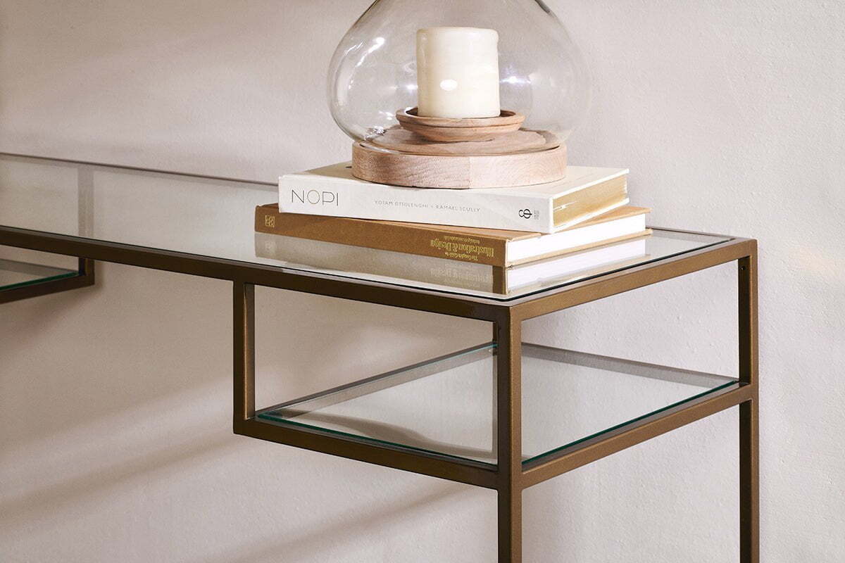 The nkuku Nakuru Iron & Glass Console Table features a bronze frame and glass top. Stacked books, including “NOPI,” sit atop it, alongside a round glass candle holder with a white pillar candle, set against a light-colored wall.