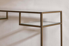 A close-up of the Nakuru Iron & Glass Console Table by nkuku shows its slim metallic bronze frame and clear glass shelves against a white wall, casting soft shadows. The minimalist design features clean, straight lines for a modern look.