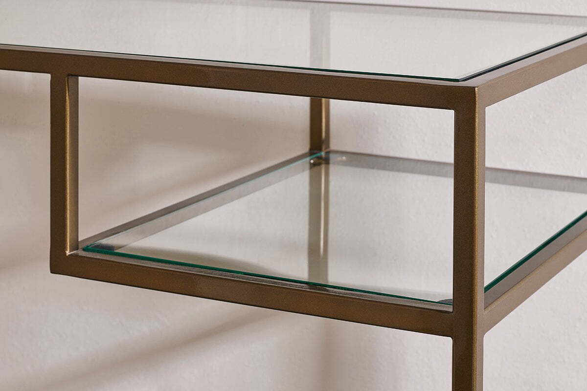 Close-up of the Nakuru Iron & Glass Console Table by nkuku, showcasing its bronze iron frame and clear glass surfaces. This contemporary hallway table features clean lines and a sleek look against a plain white wall.