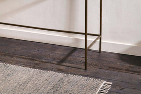 A close-up of dark wooden floorboards, a textured light gray fringed rug, white baseboards, and the slender bronze legs of the nkuku Nakuru Iron & Glass Console Table against a light-colored wall.