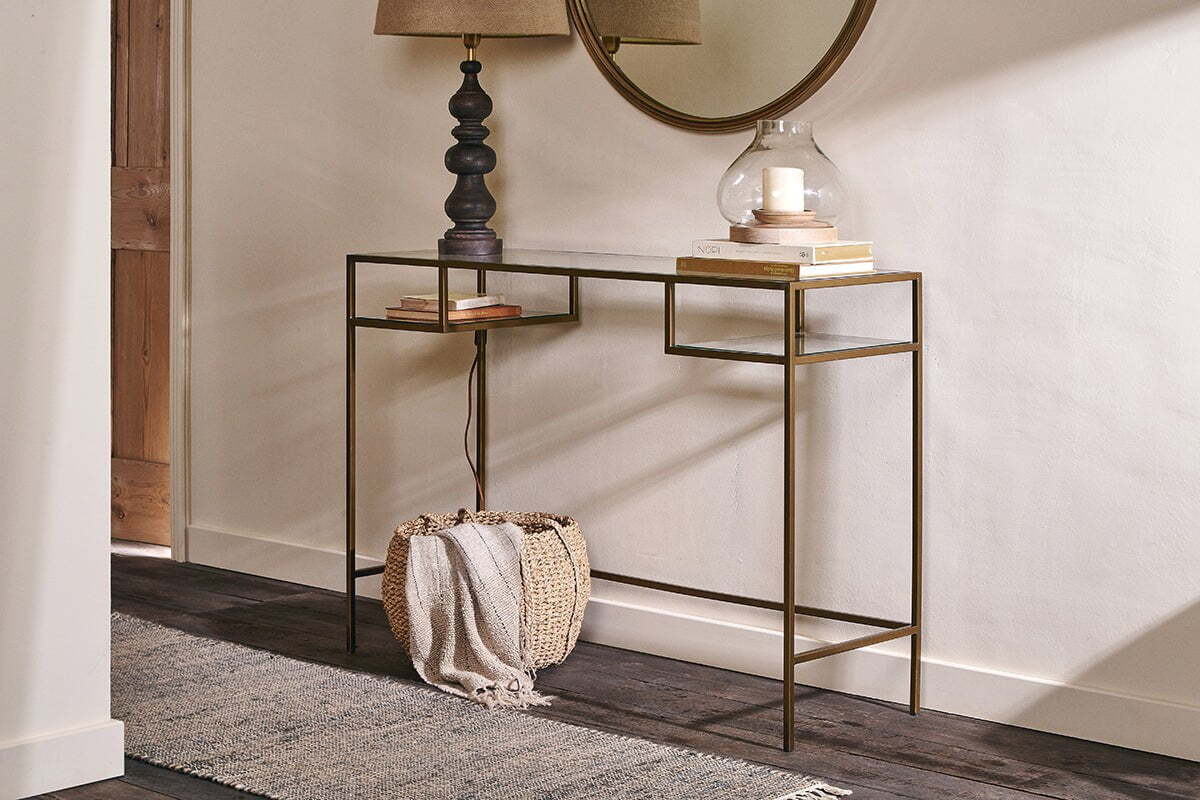 The Nakuru Iron & Glass Console Table by nkuku features a slim gold frame and glass top styled with a black lamp, glass candle holder, and books. A woven basket with a blanket sits below, while a round mirror and rug complete the look on dark wood floors.