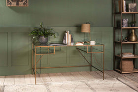 The Nakuru Iron & Glass Desk by nkuku, styled with a laptop, plant, lamp, and books, brings modern elegance to a home office set against a bold green paneled wall.