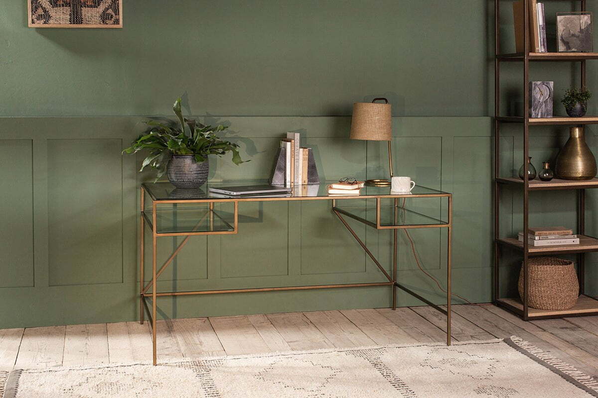 The Nakuru Iron & Glass Desk by nkuku, styled with a laptop, plant, lamp, and books, brings modern elegance to a home office set against a bold green paneled wall.