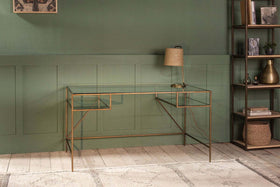 The nkuku Nakuru Iron & Glass Desk and a tan lamp stand out against a dark green paneled wall, creating an elegant home office setup. A tall shelving unit on the right displays decorative accents.