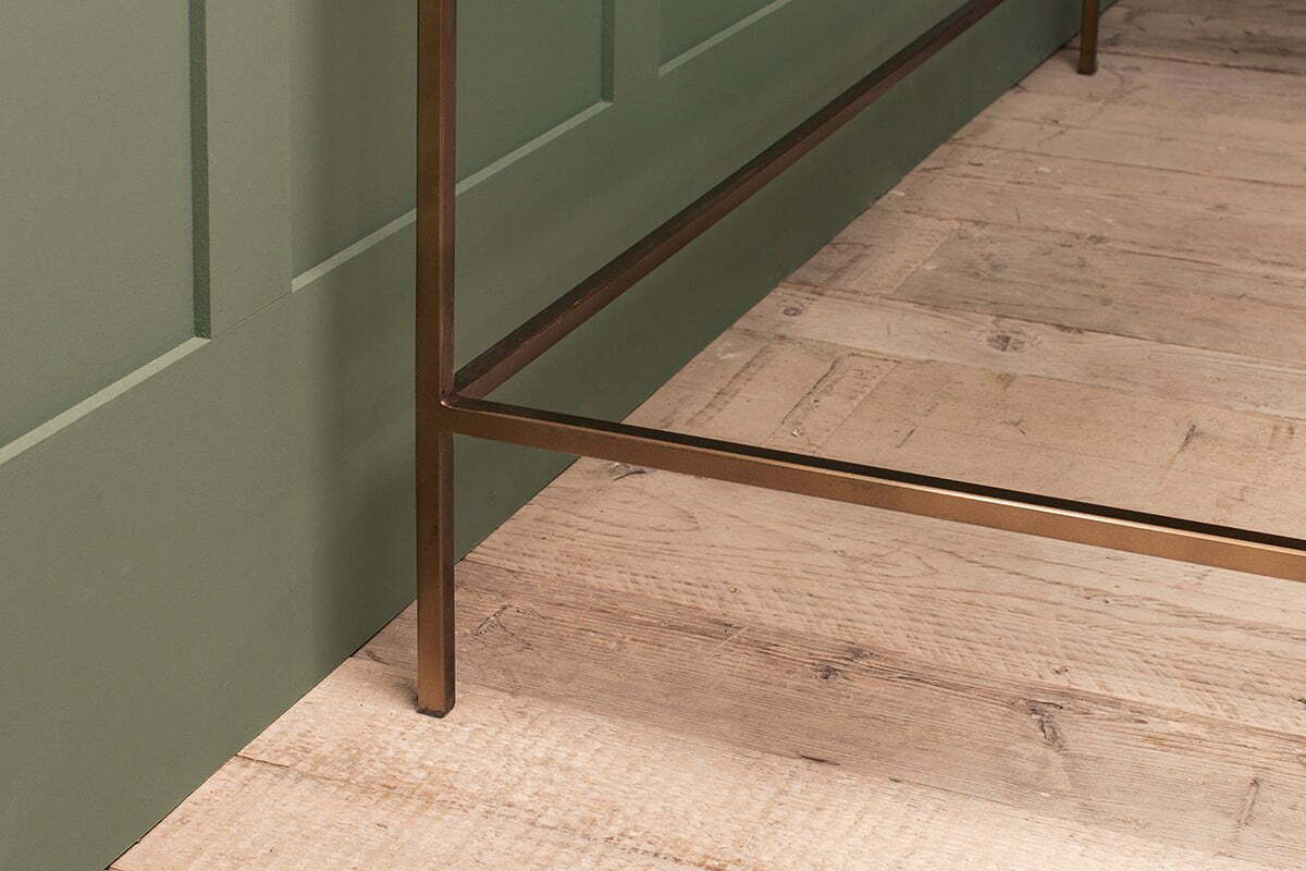 A close-up of the nkuku Nakuru Iron & Glass Desk showcases its bronze-finished metal leg on light wood flooring beside a green paneled wall. The visible wood grain and imperfections lend the scene a rustic charm.