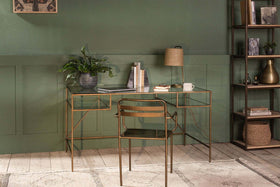 The Nakuru Iron & Glass Desk by nkuku, featuring a gold frame and glass top, holds books, a plant, lamp, and mug. Paired with a gold chair, it stands before a green paneled wall; shelves on the right display decor on wooden flooring.