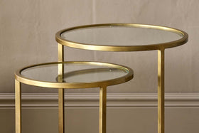 A pair of nkuku Nakuru Iron & Glass Side Table Set - Brass, featuring round glass tops and brass frames, sit elegantly against a textured wall and baseboard.