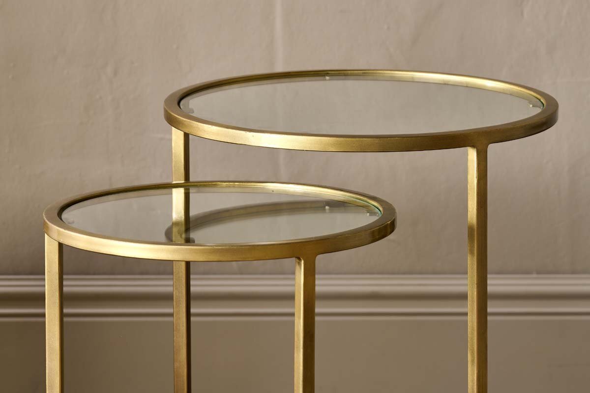 A pair of nkuku Nakuru Iron & Glass Side Table Set - Brass, featuring round glass tops and brass frames, sit elegantly against a textured wall and baseboard.
