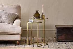 Two nested nkuku Nakuru Iron & Glass Side Tables in brass hold a glass of water, two brown vases, and a pink candle beside a beige sofa with a textured cushion, next to a dark wooden trunk on a rustic wooden floor.