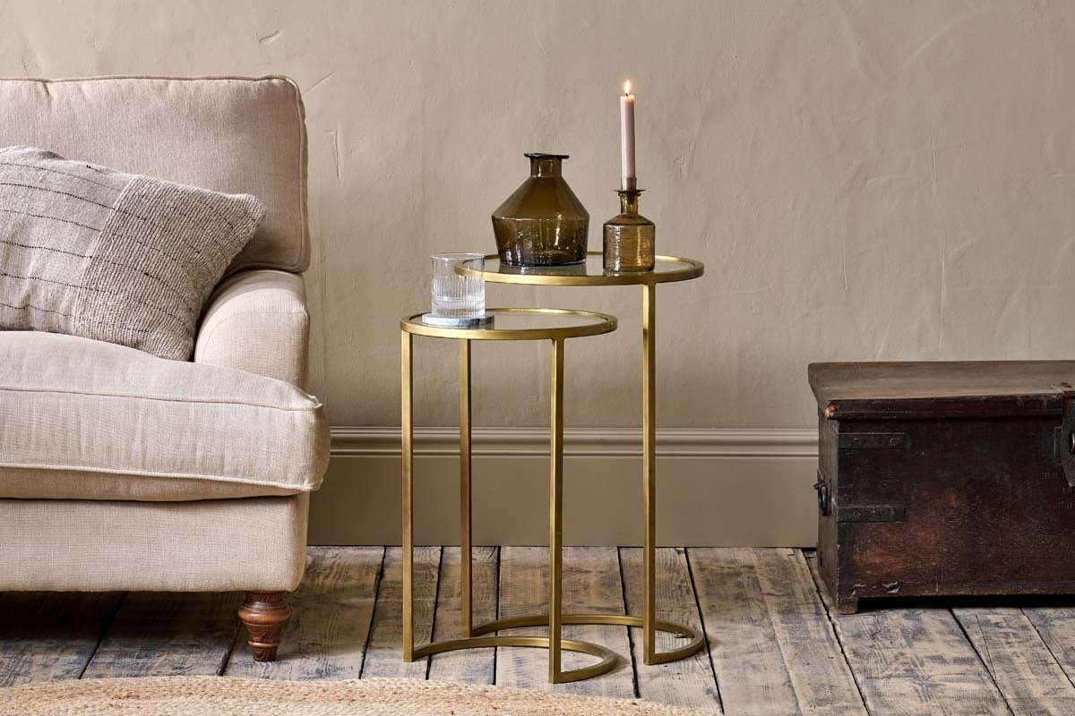 Two nested nkuku Nakuru Iron & Glass Side Tables in brass hold a glass of water, two brown vases, and a pink candle beside a beige sofa with a textured cushion, next to a dark wooden trunk on a rustic wooden floor.