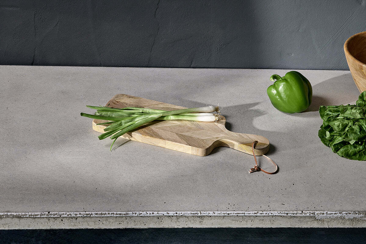 Green onions rest on the Nalbari Chopping Board - Natural by nkuku, made from sustainable mango wood, set on a gray countertop with a green bell pepper, leafy greens, a wooden bowl, and a gray wall in the background.