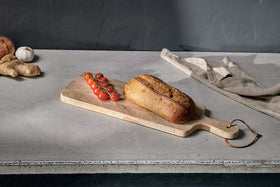 The nkuku Nalbari Chopping Board - Natural sits on a concrete countertop with rustic bread and vine cherry tomatoes. In the background, garlic, ginger, onion, and a folded beige cloth rest against a gray wall.