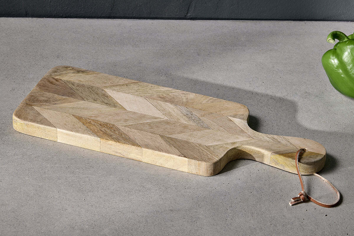 The nkuku Nalbari Chopping Board - Natural, featuring a chevron pattern and leather loop, made from sustainable mango wood, rests on a gray countertop with a green bell pepper visible against the dark gray wall.